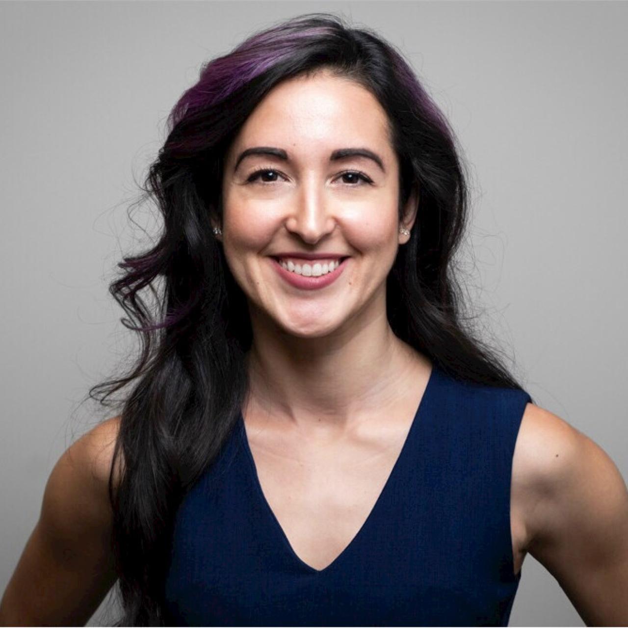 Amelia smiling into camera with blue v-neck blouse against gray background.