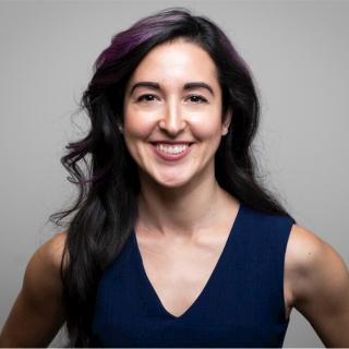 Amelia smiling into camera with blue v-neck blouse against gray background.